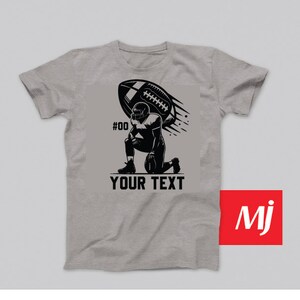 K&ouml;nnte beinhalten: Ein graues T-Shirt mit einer schwarzen Illustration eines Fussballspielers, der kniet und einen Fussball in der Luft h&auml;lt. Der Text "#00" und "YOUR TEXT" ist auf dem T-Shirt aufgedruckt.