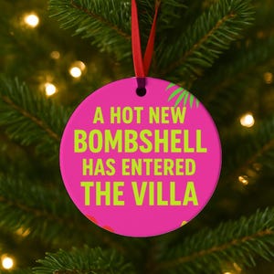 May include: A round, pink Christmas ornament with the text "A HOT NEW BOMBSHELL HAS ENTERED THE VILLA" in bright green. The ornament hangs from a red ribbon against a backdrop of a Christmas tree.