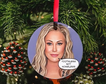 Peut inclure: Une boule de Noël ronde représentant un portrait de femme en dessin animé avec des cheveux blonds. La boule a un fond violet et une bulle de dialogue avec le texte "Toothless Not Homeless".