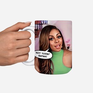 May include: White ceramic mug with a colorful image of a woman with brown hair and a green top. The woman is looking at the camera and has a speech bubble above her head that says "Not Today Satan!"