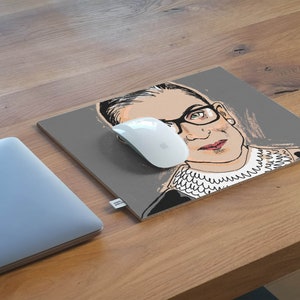 May include: A gray mousepad with a black and white illustration of Ruth Bader Ginsburg. The illustration shows her wearing a black robe and a white lace collar. The mousepad is on a wooden surface.