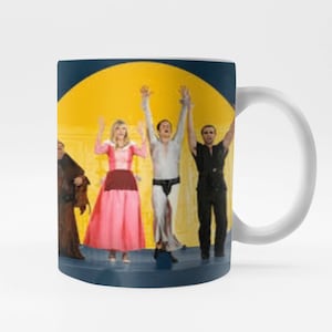 May include: A white ceramic mug with a blue rim and handle. The mug features a colorful image of a group of people singing and dancing on a stage with a yellow sun behind them.