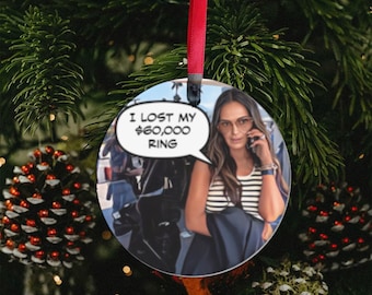 Peut inclure: Une boule de Noël ronde avec un fond blanc et une image de bande dessinée d'une femme aux longs cheveux bruns portant une veste noire et une chemise rayée. La femme tient un téléphone portable à son oreille et une bulle de dialogue au-dessus de sa tête dit "J'ai perdu ma bague à 60 000 €."