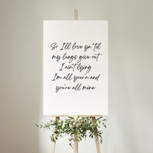 May include: A white canvas sign with black calligraphy text that reads "So I'll love ya til my lungs give out I ain't lying I'm all your'n and you're all mine". The sign is on an easel with green leaves and white flowers.
