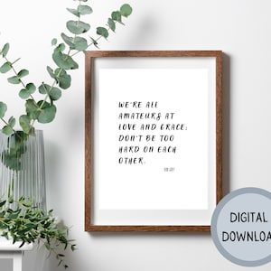 May include: A framed print with a white background and black text that reads "We're all amateurs at love and grace: Don't be too hard on each other." The text is attributed to Mob Goff. The frame is brown wood.