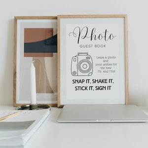 Peut inclure: Un panneau encadré "Photo Guest Book" avec une illustration d'appareil photo et le texte "Snap it, Shake it, Stick it, Sign it." Un ordinateur portable, une bougie et une œuvre d'art sont sur une surface blanche.