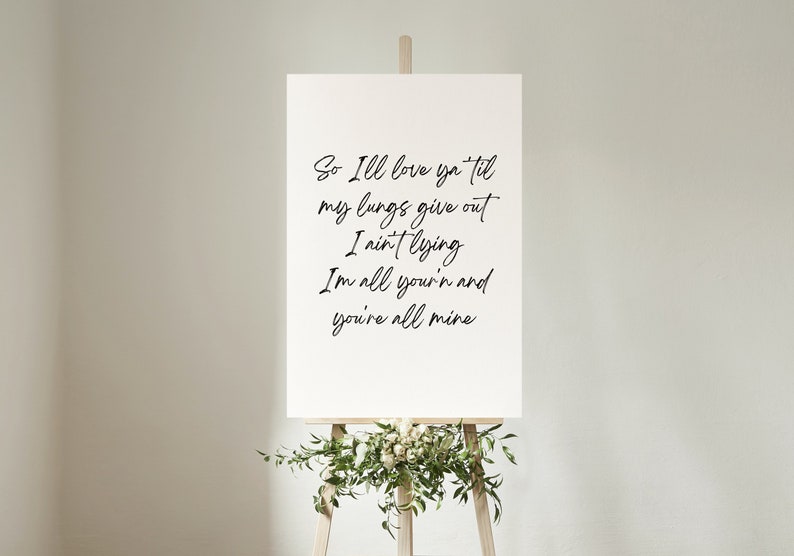 May include: A white canvas sign with black calligraphy text that reads "So I'll love ya til my lungs give out I ain't lying I'm all your'n and you're all mine". The sign is on an easel with green leaves and white flowers.