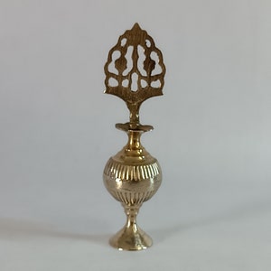 Puede incluir: Frasco de perfume dorado con una parte superior decorativa calada. El frasco tiene un cuerpo bulboso con estrías verticales y una base delgada y acampanada. La parte superior presenta un diseño ornamentado y estilizado.