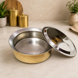 May include: A hammered metal serving bowl with a gold-colored exterior and a silver-colored interior. The lid is partially open, revealing the inside of the bowl. The bowl is round and sits on a light-colored surface.