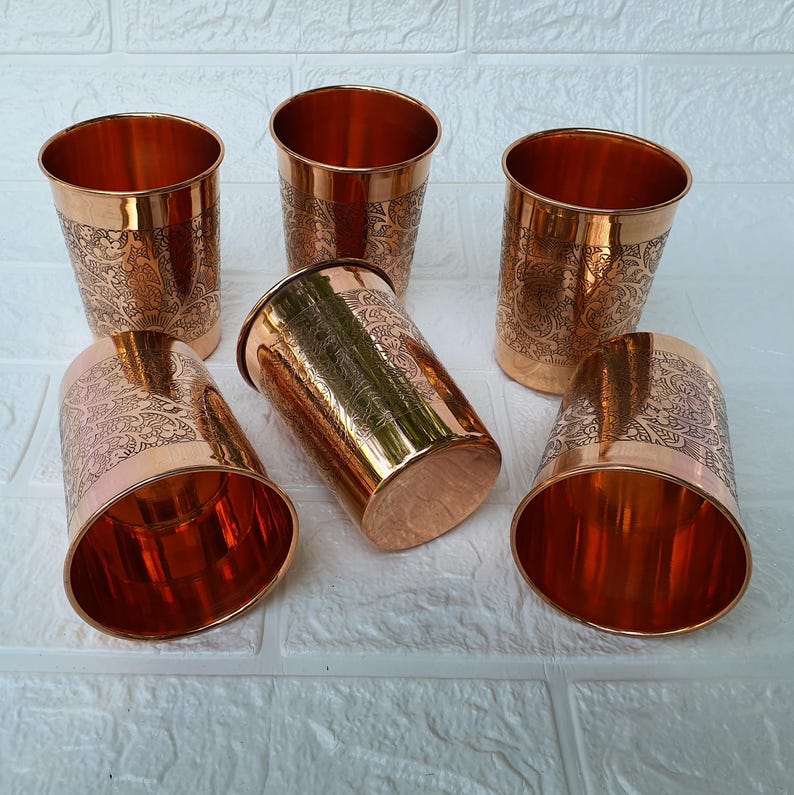 May include: Six copper-colored drinking glasses with etched floral designs. The glasses are cylindrical with a slightly flared rim. The interior of the glasses is a bright copper color, and the exterior has a darker, more antique finish.