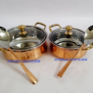Può includere: Due ciotole da portata in rame martellato con coperchi e mestoli abbinati. Le ciotole hanno un interno in acciaio inossidabile e manici dorati.