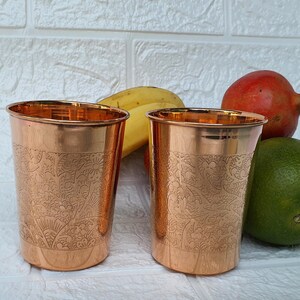 May include: Two copper-colored drinking glasses with etched floral patterns. The glasses have a shiny finish and a slightly flared rim. The background includes a banana, a pomegranate, and a lime.