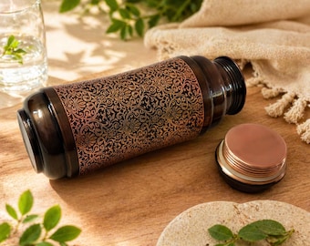 Op de afbeelding: Een donkerbruine waterfles met een koperkleurig bloemmotief. De fles ligt op een houten oppervlak met het deksel ernaast. Een glas water en groen zijn op de achtergrond te zien. De fles is ongeveer 25 cm lang.