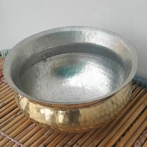 May include: A hammered metal bowl with a brass base and a silver-coloured rim. The bowl has a wide, open design and sits on a bamboo mat. The brass base has a textured, dimpled surface.