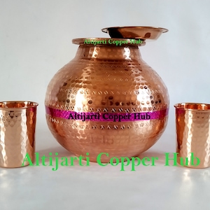 May include: A copper water pot with a hammered texture, a small plate, and two matching drinking glasses. The pot has a wide base and a narrow neck. A pink band wraps around the middle of the pot.