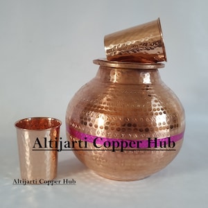 May include: A copper water pot with a lid, a small cup on top, and a drinking glass. The copper items have a hammered texture and a pink band around the pot. The text "Altijarti Copper Hub" is visible.
