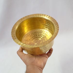 Op de afbeelding: Een gehamerde messing kom met een gouden afwerking. De kom heeft een afgeronde vorm met een getextureerd oppervlak en wordt in een hand gehouden tegen een witte achtergrond. De binnen- en buitenkant van de kom zijn goudkleurig.