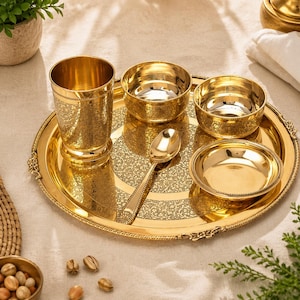 May include: A gold-coloured serving set with detailed floral patterns. The set includes a round tray, a tall glass, three bowls, and a spoon. The items are arranged on a light-coloured surface, suggesting a dining or serving context.