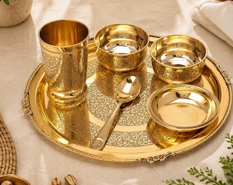 Puede incluir: Un juego de servir dorado con intrincados diseños florales. El conjunto incluye una bandeja redonda, un vaso alto, tres cuencos y una cuchara. Los artículos están dispuestos sobre una superficie clara, sugiriendo un contexto de comedor o servicio.