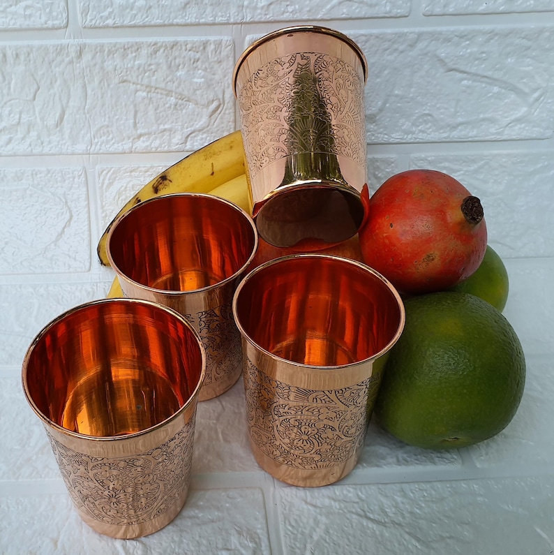 May include: Four copper-colored drinking glasses with etched floral designs, alongside a larger glass. A banana, a pomegranate, and two limes are also visible. The glasses have a shiny, reflective interior.