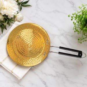 May include: A round, gold-coloured metal pan with a textured, hammered surface featuring a concentric circular pattern. The pan includes a long, silver-toned metal handle with two black plastic grips and a loop at the end for convenient hanging.