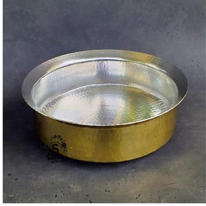 May include: A hammered brass bowl with a silver interior. The bowl features a wide, rounded rim and a slightly textured surface. The exterior is a warm gold colour, and the interior has a reflective, polished finish. The bowl has a small logo on the side.