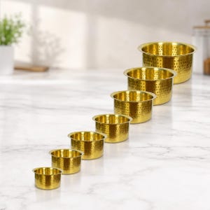 Op de afbeelding: Een set van zes goudkleurige metalen kommen van verschillende maten, gerangschikt in aflopende volgorde. Elke kom heeft een afgeronde rand en is bedekt met kleine, gelijkmatig verdeelde perforaties. De kommen staan op een wit marmeren oppervlak.