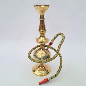 May include: A gold-colored hookah with intricate etched designs. The hookah has a bulbous base, a slender central column, and a flared top. A colorful hose with red and orange accents is attached. The hookah is a decorative item.