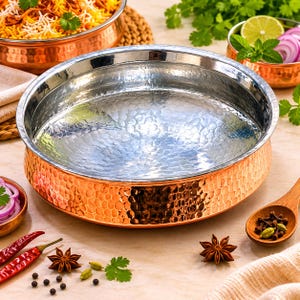May include: A large, hammered copper serving dish with a silver interior. The dish is round with a wide rim and sits on a light-colored surface. Other copper bowls and ingredients like spices and herbs surround the dish.