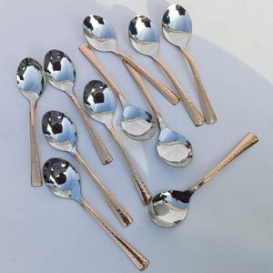 May include: An assortment of polished silver spoons with copper-toned, hammered handles. The spoons are arranged on a light surface, reflecting the surroundings. These spoons are designed for serving.