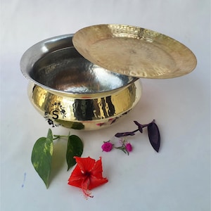 May include: A hammered brass serving bowl with a lid. The bowl has a silver interior and a gold exterior. The bowl is sitting on a white surface with flowers and leaves.