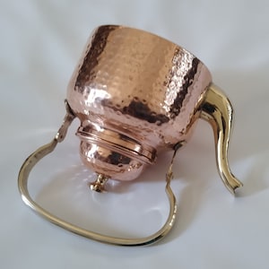 Può includere: Una teiera in metallo color rame con una trama martellata. Presenta un manico color oro e un manico curvo color argento. La teiera ha un coperchio e una piccola base decorativa. Il design è vintage e decorativo.