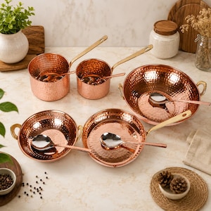 May include: A set of five hammered copper-toned cookware pieces including two saucepans with long handles, two round bowls with side handles, and one shallow pan with a long handle. Each piece contains a polished silver-toned metal spoon.