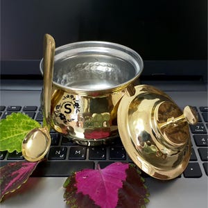 Puede incluir: Un azucarero de metal dorado con tapa y cuchara a juego. El cuenco tiene una textura martillada y un pequeño logotipo con la letra "S". La tapa está parcialmente abierta, revelando el interior del cuenco. La cuchara descansa junto al cuenco.