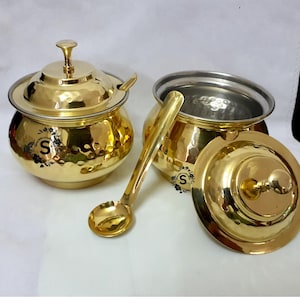 May include: Two gold-coloured, hammered-finish serving bowls with lids and a serving spoon. One bowl has its lid on, and the other has its lid off. The bowls have a silver interior. The spoon is resting on the side of the bowl with the lid on.