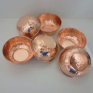May include: Six copper-coloured bowls with a hammered texture. The bowls are round with a slightly flared rim. The interior of each bowl has a decorative pattern. The bowls are arranged on a white surface.