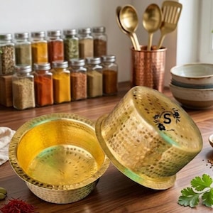 May include: Two hammered gold-colored bowls with a textured surface. The bowls have a wide, shallow design and a bright interior. The bowls are next to a collection of spices in glass jars, and a copper utensil holder.