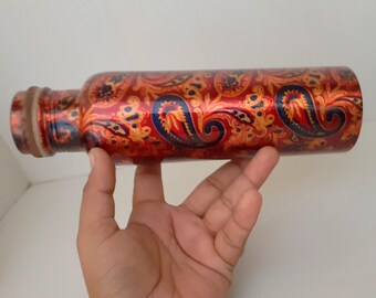 May include: A decorative water bottle with a paisley pattern in red, gold, and blue. The bottle is cylindrical with a narrow neck and a wooden-looking cap. The bottle is held by a hand.