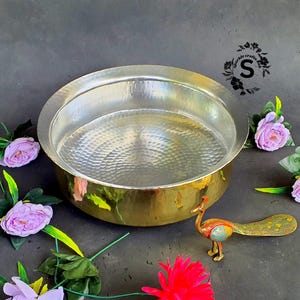 Puede incluir: Un cuenco de metal martillado con base de latón e interior plateado. Una pequeña figura de pavo real de colores está junto al cuenco. El fondo es gris oscuro, con flores rosas y moradas.