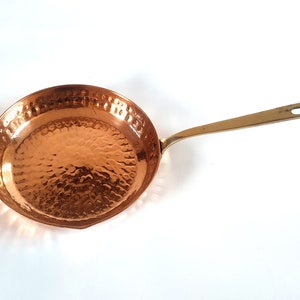 Puede incluir: Sartén de color cobre con una textura martillada en la superficie de cocción. El mango, de color latón, es largo y tiene un agujero para colgar. Diseñada para cocinar.