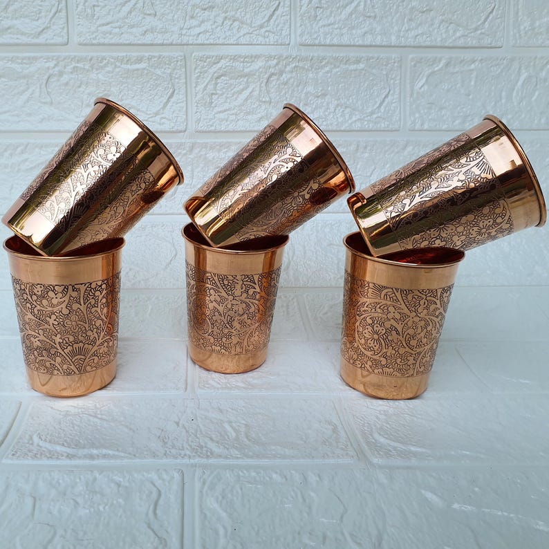 May include: Six copper-colored drinking glasses with intricate floral patterns. The glasses are arranged in a stacked configuration, with three upright and three tilted. The copper has a shiny finish, reflecting light.