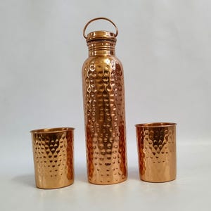 Puede incluir: Un juego de tres vasos para beber de cobre con textura martillada. El juego incluye una botella alta con tapa y dos vasos más cortos.