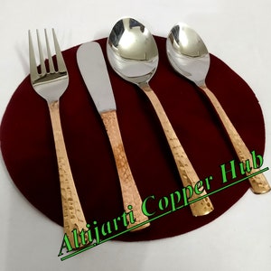 Puede incluir: Un juego de cuatro cubiertos: un tenedor, un cuchillo de mantequilla y dos cucharas. La cubertería tiene cabezas de acero inoxidable y mangos martillados de color cobre. El juego se muestra sobre una superficie circular roja oscura.
