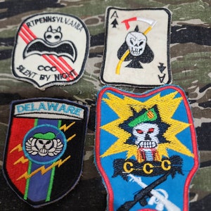 May include: Four embroidered patches with military themes. The top left patch is white with red stripes and black bat, with the text "RT.PENNSYLVANIA. SILENT BY NIGHT". The top right patch is white with a black spade, a skull, and a scythe, with the text "A". The bottom left patch is red, blue, green, and black with the text "DELAWARE" and a skull with wings. The bottom right patch is blue, yellow, red, and white with a skull, a rifle, and the text "CCCC".