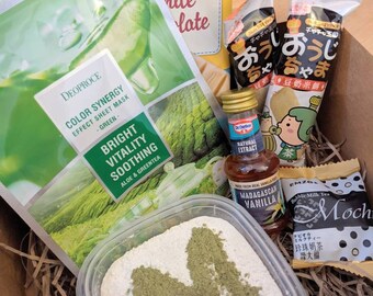 May include: A brown cardboard box filled with various items, including a green face mask, a white chocolate bar, a small bottle of vanilla extract, a package of mochi, and two packages of Japanese snacks. The box is filled with brown shredded paper.