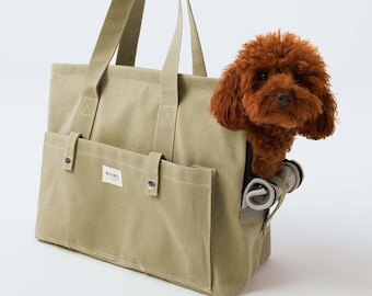 May include: A light olive green canvas tote bag with a small brown dog peeking out. The bag has two exterior pockets and long handles. The word "BRUNO" is printed on a small white label on the front of the bag.