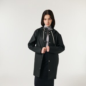 May include: A black, hooded raincoat with a button-up front and white drawstrings. The model is wearing a gray turtleneck, a dark gray skirt, and brown loafers. The raincoat is a classic outerwear piece.