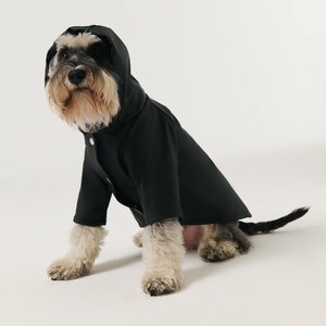 May include: A small, black dog wearing a black raincoat with a hood. The dog is sitting on a white background.