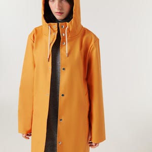 May include: Mustard-colored hooded raincoat with a drawstring hood and snap closures. The long-sleeved coat is worn over a gray turtleneck. The coat is a solid color and has a simple, classic design.