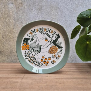 Può includere: Un piatto in ceramica verde con un bordo bianco presenta un'illustrazione stilizzata di una persona che raccoglie frutta da un albero. Il design include frutta arancione e verde, foglie e viti.